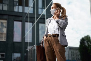 Koronavirüs sırasında güvenli bir şekilde çalışıyor. Kendine güveni tam bir iş kadını koruyucu yüz maskesi takıyor, dışarıda ofis binasının önünde beklerken cep telefonuyla konuşuyor.