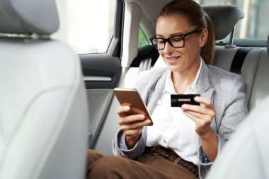 Çevrimiçi ödeme. Gülümseyen iş kadını gözlüklü, akıllı telefonu ve kredi kartıyla arabada arka koltukta otururken bir şeyler alıyor.