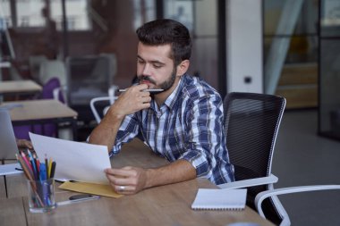 Kafkasyalı genç bir işadamı ya da erkek ofis çalışanı iş yerindeki bazı belgeleri analiz ediyor, modern çalışma alanındaki iş yerinde oturuyor.