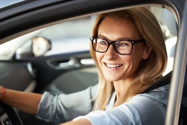Siap untuk perjalanan bisnis. Potret seorang wanita bisnis Kaukasia yang bahagia mengenakan kacamata duduk di mobil modernnya, melihat kamera dan tersenyum — Stok Foto