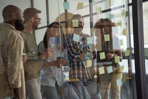 Young multiracial business team, group of creative people looking at sticky notes on glass board and discussing something, working together in modern office