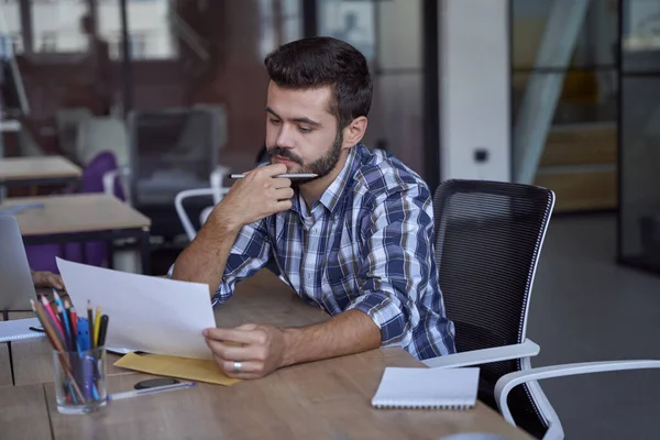Kafkasyalı genç bir işadamı ya da erkek ofis çalışanı iş yerindeki bazı belgeleri analiz ediyor, modern çalışma alanındaki iş yerinde oturuyor.