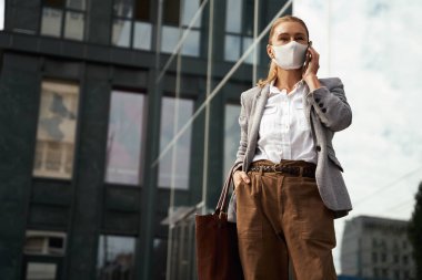 Covid 19 sırasında iş. Kendine güveni tam bir iş kadını koruyucu yüz maskesi takıyor, dışarıda ofis binasının önünde beklerken cep telefonuyla konuşuyor.