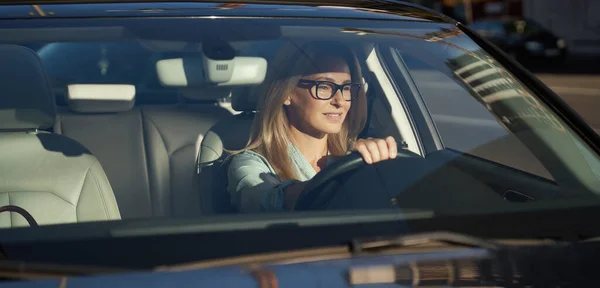 Šťastná atraktivní běloška s brýlemi, která ráno řídí své moderní auto po městě, dívá se na silnici a usmívá se — Stock fotografie