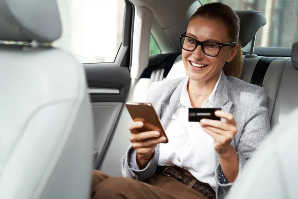 Wanita bisnis yang ceria memakai kacamata menggunakan smartphone dan kartu kreditnya untuk membeli sesuatu dan tersenyum sambil duduk di kursi belakang mobil — Stok Foto