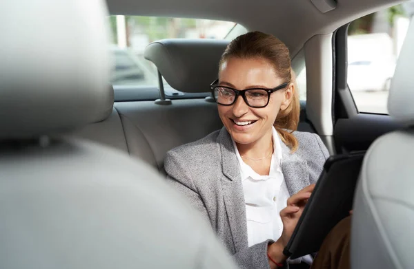 Bekerja dalam perjalanan ke kantor. Wanita bisnis yang sukses mengenakan kacamata menggunakan tablet digital dan tersenyum sambil duduk di kursi belakang mobil — Stok Foto