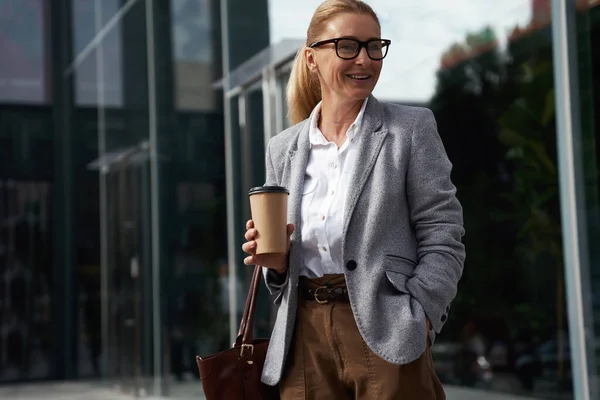 Istirahat kopi. Wanita bisnis yang gembira memakai kacamata klasik sambil memegang secangkir kopi dan tersenyum sambil berdiri di depan gedung kantor kaca di luar ruangan — Stok Foto