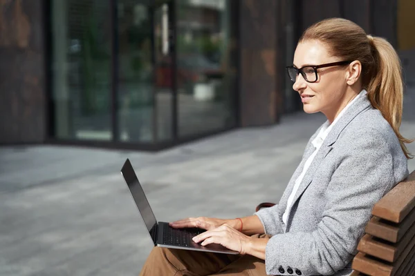 Bekerja online. Tampilan samping dari seorang pengusaha wanita sukses yang bekerja pada laptop sambil duduk di bangku cadangan di luar gedung kantor — Stok Foto