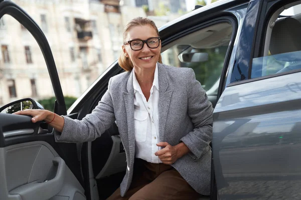 Przyjeżdżam do biura. Portret atrakcyjnej i szczęśliwej biznesmenki w okularach wysiadającej z nowoczesnego samochodu i uśmiechniętej — Zdjęcie stockowe