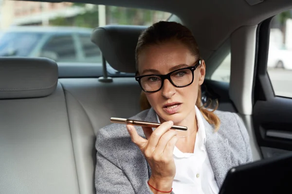 Wielozadaniowość. Portret pewnej siebie kobiety biznesowej korzystającej z tabletu cyfrowego i smartfona, nagrywającej wiadomość głosową siedząc na tylnym siedzeniu samochodu — Zdjęcie stockowe