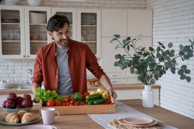 Vejetaryenlik kavramı. Çeşitli taze sebzelerle dolu sepet tutan ve evde sağlıklı yemekler pişiren mutlu genç bir adam.