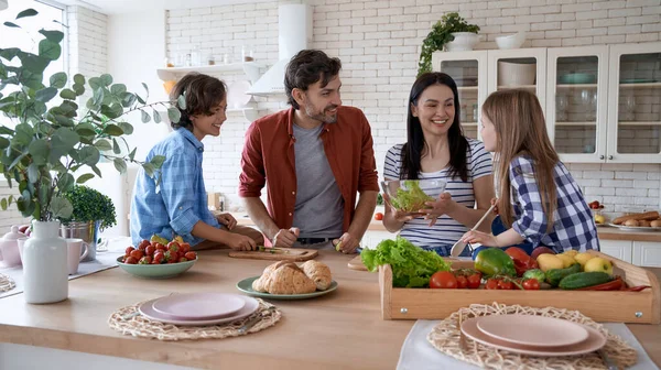 Family day at home. Mother, father and two little excited kids cooking together in the modern kitchen at home. They are preparing a salad, talking and smiling — Stock Photo, Image