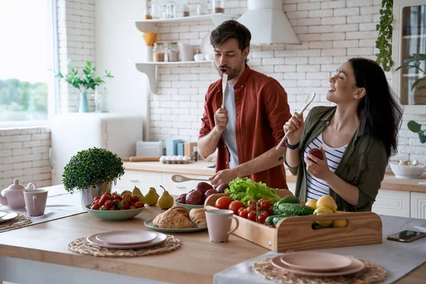 Young happy family couple dancing, singing and having fun while preparing healthy food in the modern kitchen at home. Fresh fruits and vegetables on the wooden table — Stock Photo, Image