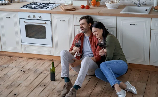 Romantic evening at home. Young happy romantic couple, wife and husband sitting on the floor in the modern kitchen, drinking wine and hugging