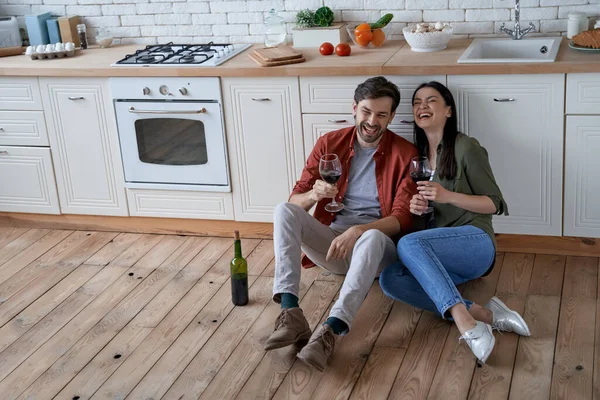 Spending time at home. Young happy romantic couple, wife and husband sitting on the floor in the modern kitchen, drinking wine and talking after cooking together