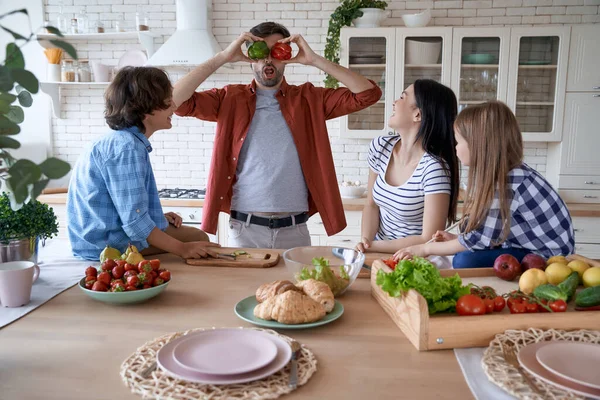 Funny dad playing with sweet pepper while cooking with his family together in the modern kitchen at home. Mother, father and two little excited preparing a salad — Stock Photo, Image