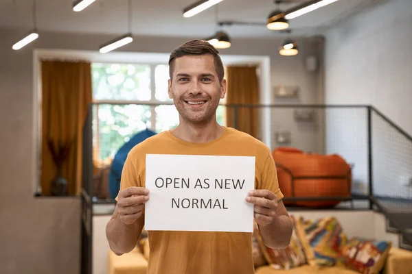 We are open. Young happy man, office worker showing paper with text OPEN AS NEW NORMAL and smiling at camera, standing in the modern coworking space