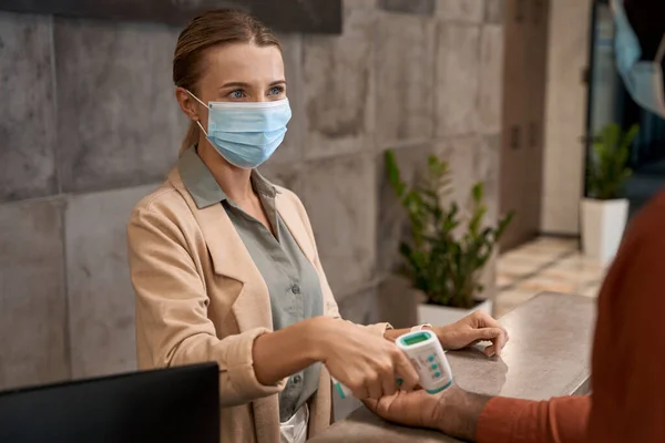 Mujer joven con mascarilla médica de pie en la recepción y el uso de termómetro infrarrojo para comprobar la temperatura de las personas, los trabajadores de oficina — Foto de Stock