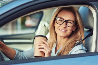 Modern arabasının direksiyonunda oturan, elinde kahveyle kameraya bakan, gülümseyen olgun bir kadının portresi.