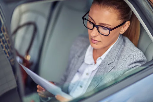 Concentrated middle aged businesswoman wearing eyeglasses reading documents while sitting on back seat in the car, preparing for a meeting