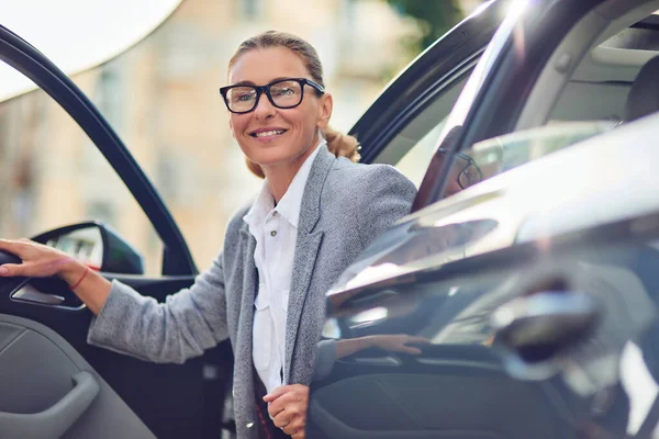 Wanita bisnis paruh baya yang positif dan sukses dalam pakaian klasik keluar dari mobil modernnya dan tersenyum, pergi untuk pertemuan bisnis — Stok Foto