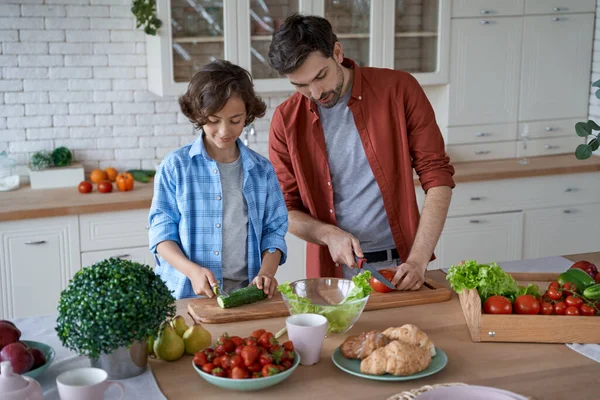 Birlikte yemek hazırlıyoruz. Genç baba oğluna taze sebze kesmeyi öğretiyor, salata hazırlıyor, evde modern mutfakta duruyor. — Stok fotoğraf