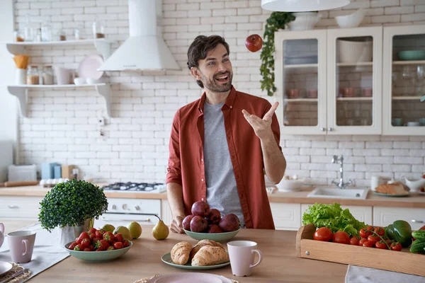 每天早上吃苹果。年轻快乐的大胡子男人穿着休闲装，站在现代化厨房里，吐着一个红色的苹果，微笑着 — 图库照片