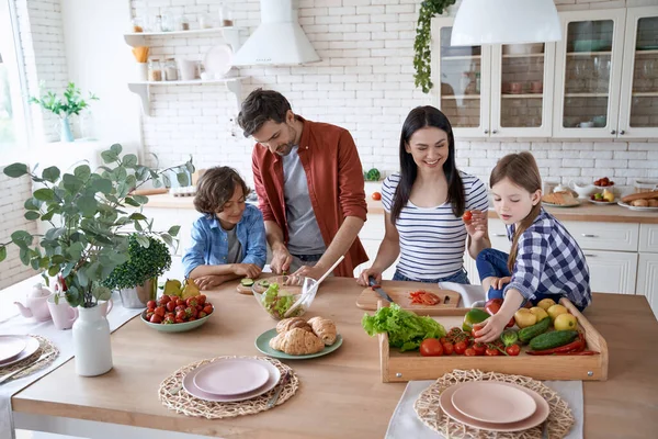 Salata hazırlıyorum. Genç ve güzel bir aile evdeki modern mutfakta birlikte yemek pişiriyor. Anne ve baba iki küçük çocuğa taze sebze kesmeyi öğretiyor. — Stok fotoğraf