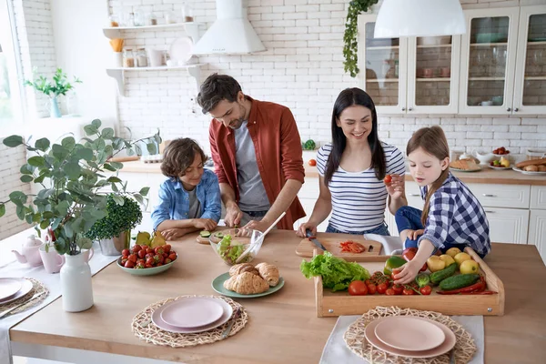Menyiapkan salad. Keluarga muda yang cantik memasak bersama di dapur modern di rumah. Ibu dan ayah mengajari dua anak kecil cara memotong sayuran segar Stok Lukisan  