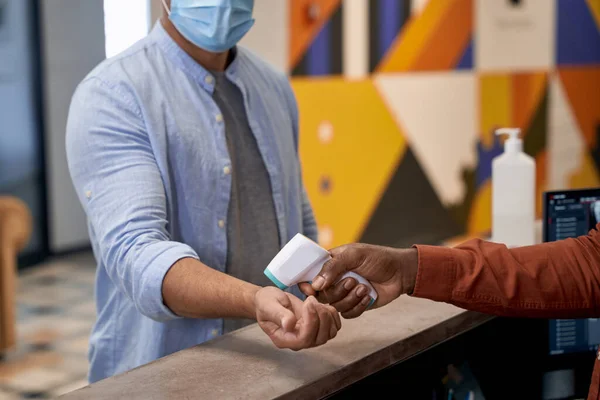 Detección de temperatura en el trabajo, recepcionista africano masculino usando máscara médica usando termómetro infrarrojo para medir la temperatura corporal del trabajador de oficina — Foto de Stock