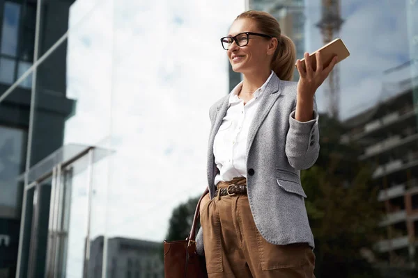 Happy successful business woman wearing eyeglass and classic wear holding mobile phone and smile while standing against office building outdoor — Stok Foto