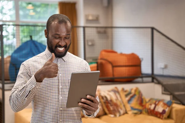 Potret seorang pemuda pekerja kantor laki-laki afrika yang bahagia menggunakan tablet digital, tersenyum dan menunjukkan jempol saat bekerja di kantor modern — Stok Foto