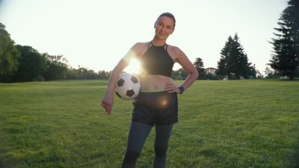 Voetbalspeler. Portret van een mooie en sportieve vrouw die voetbal in de hand houdt, naar de camera kijkt en glimlacht terwijl ze in een groen veld staat — Stockvideo