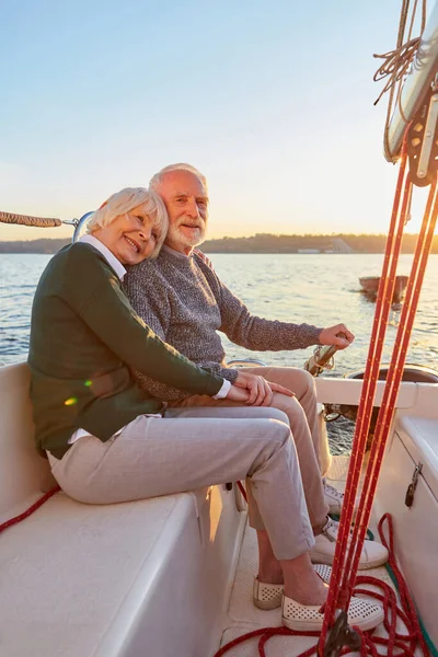 Full leghth of happy senior couple, elderly man and woman holding hands, hugging and spending time together while sitting on the side of yacht deck floating in sea on a sunny day