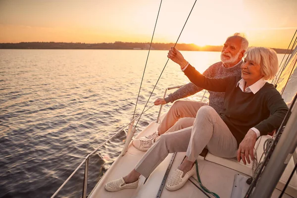 Enjoying luxury life. Beautiful happy senior couple in love relaxing on the side of sailboat or yacht deck floating in sea at sunset, looking at amazing evening view — Stock Photo, Image
