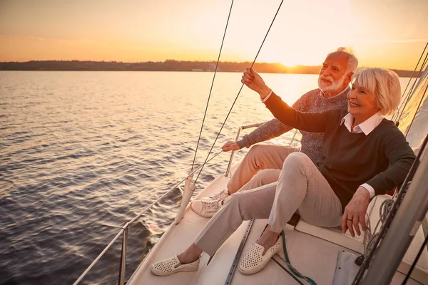 A desfrutar da vida de luxo. Casal sênior feliz bonito no amor que relaxa no lado do veleiro ou do convés do iate que flutua no mar no por do sol, olhando para a vista surpreendente da noite — Fotografia de Stock