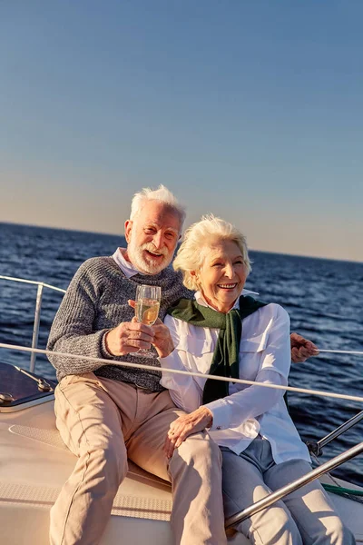 Cheers. Happy senior family couple drinking wine or champagne and smiling while celebrating wedding anniversary on a sailboat or yacht deck floating in a calm blue sea