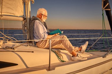 Boş zaman ve rahatlama. Günbatımında ya da gündoğumunda sakin mavi denizde yüzen yelkenli ya da yat güvertesinde otururken kıdemli adam kitap okuyor.