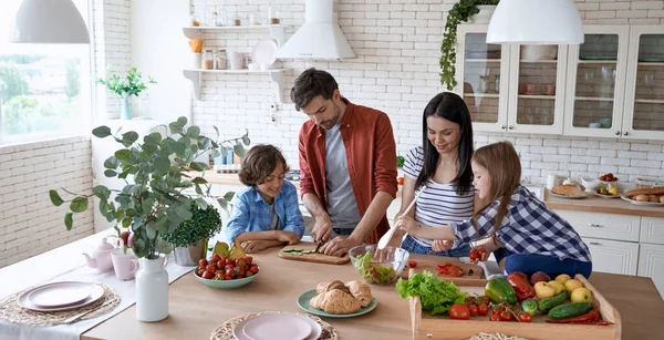 Sağlıklı beslenme. Genç ve güzel bir aile evdeki modern mutfakta birlikte yemek pişiriyor. Anne ve baba iki küçük çocuğa taze sebze kesmeyi öğretiyor.
