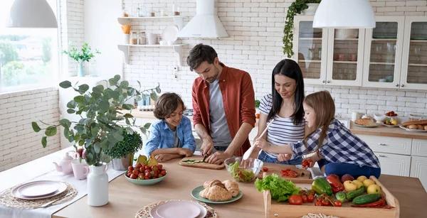 Healthy eating. Young beautiful family cooking together in the modern kitchen at home. Mother and father teaching two little kids how to cut fresh vegetables