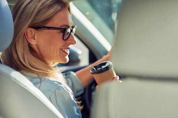 Pandangan belakang dari seorang wanita usia menengah yang cantik mengenakan kacamata duduk di belakang kemudi di dalam mobil, minum kopi dan tersenyum — Stok Foto