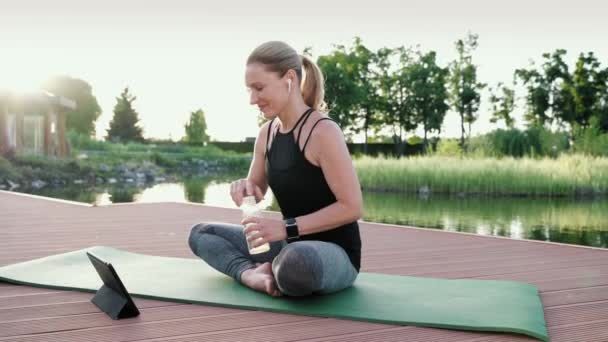 Glückliche Bloggerin, Yogalehrerin, die auf einer Matte im Park in der Nähe des schönen Sees sitzt, Wasser trinkt und nach dem Yoga-Kurs online entspannen kann. Digitales Tablet anschauen und lächeln — Stockvideo