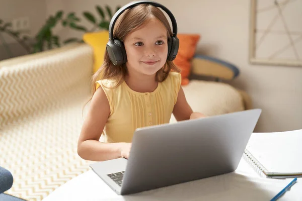 Learning online. Cute and smart little caucasian girl in headphones watching video lesson on laptop while sitting at the table in her room