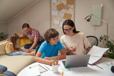 Anne ve oğul okul ödevlerini birlikte yaparken baba ve kız kanepede oturup arka planda bulmacalar çözüyorlar.