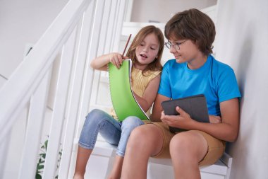 Evde eğitim. Küçük kız ve erkek kardeş evde merdivenlerde oturup ödevlerini yapıyorlar, dijital tablet kullanıyorlar.