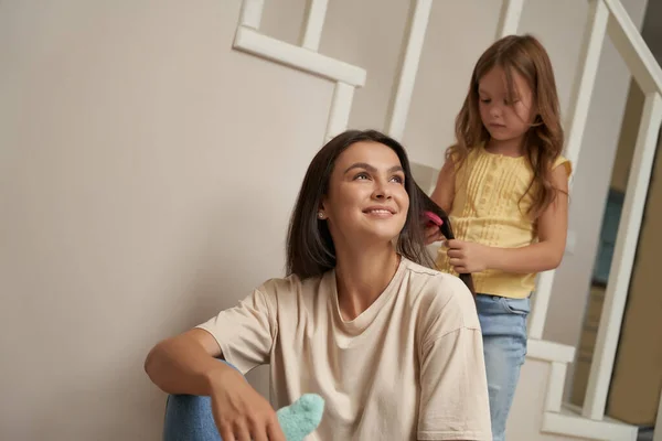 Linda niña peinando el pelo largo de su hermosa madre sonriente, mamá e hija pasando tiempo juntos en casa — Foto de Stock