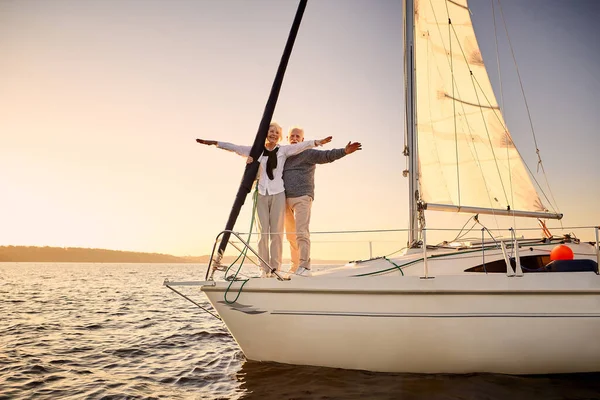 Full length of happy senior couple stretching hands out against the sky, enjoying amazing sunset and sailing together while standing on the side of yacht deck floating in the sea