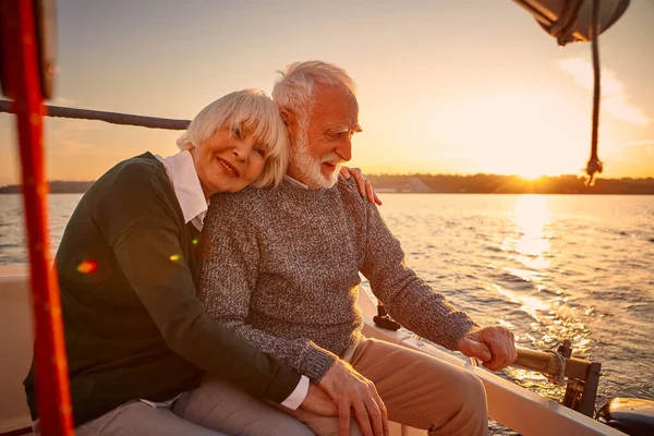 Enjoying sailing at sunset. Happy senior couple, elderly man and woman holding hands, hugging and relaxing while sitting on the side of yacht deck floating in the sea