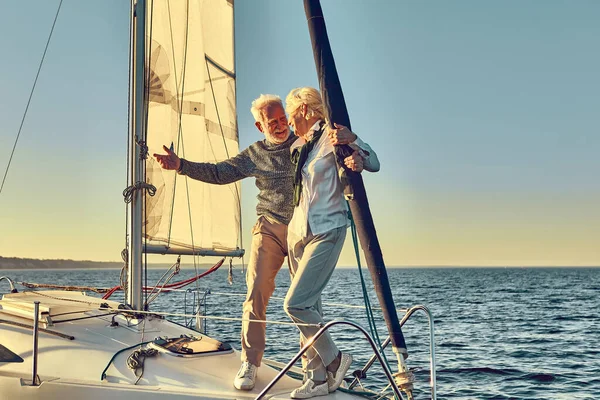 Happy senior man pointing at the horizon while standing with his wife on the side of a sail boat or yacht deck floating in a calm blue sea