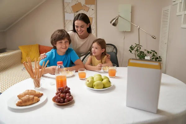 Retrato de la joven hermosa familia caucásica, madre y sus dos pequeños niños lindos sentados en la mesa de la cocina en casa y desayunando juntos — Foto de Stock
